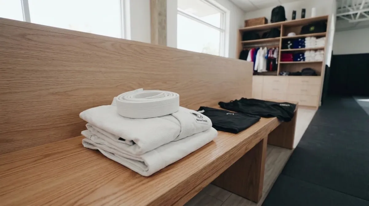 A white folded gi beside a black rash guard and shorts on a wooden bench in a bright academy