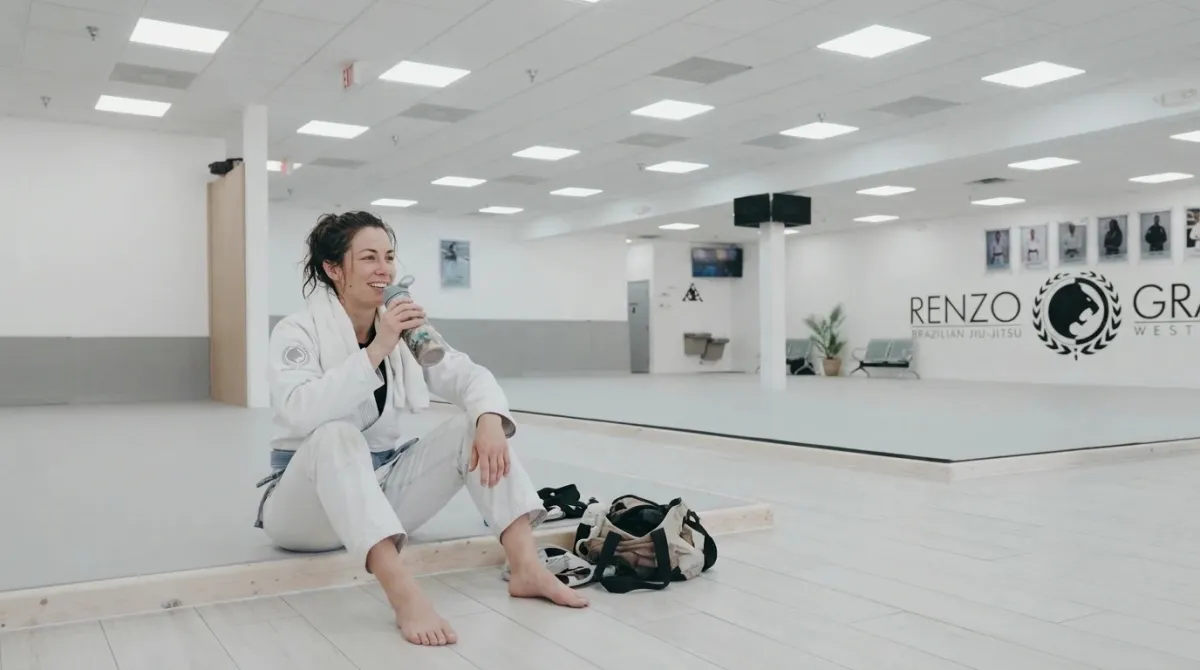 A woman with a towel on her shoulders drinking water on the edge of the mat after class