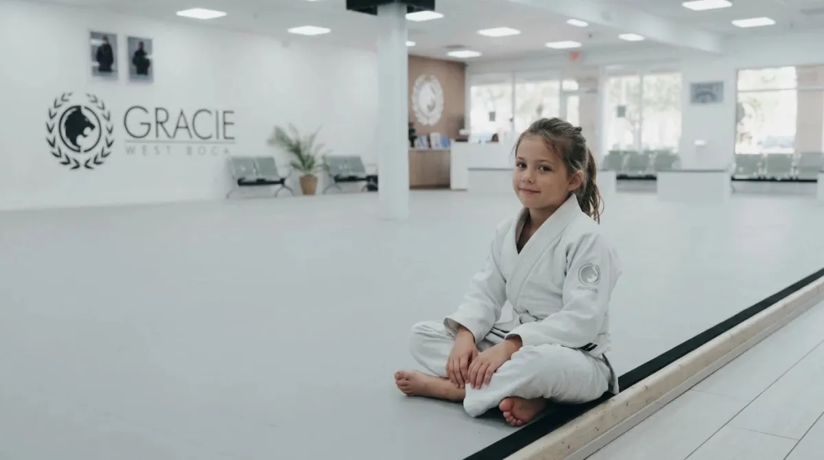 A young student in a white gi sitting quietly on the edge of a gray mat with a small smile